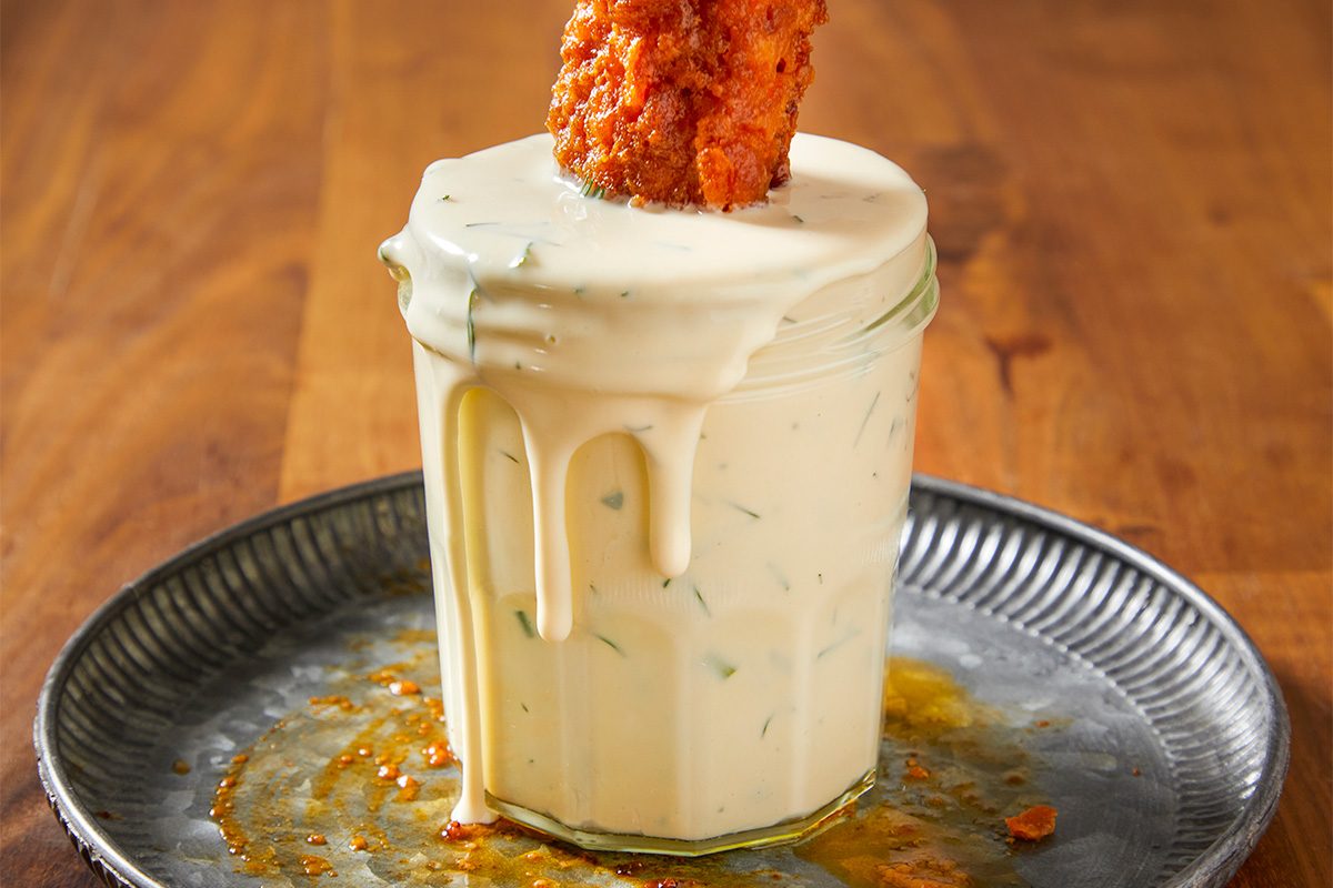 A horizontal image shows a crispy piece of fried chicken being dipped into a jar of thick ranch dressing with sauce dripping down the sides.