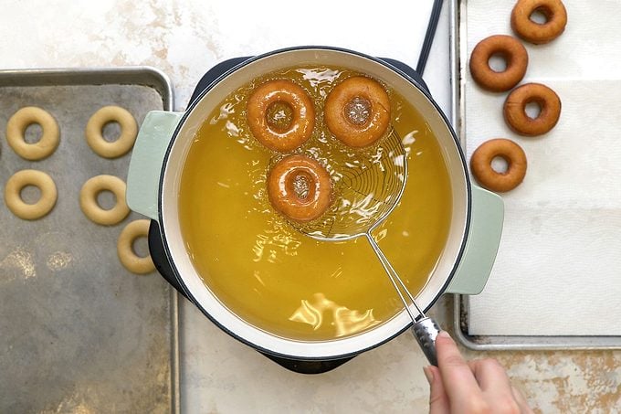 Frying doughnuts in hot oil in a large skillet with uncooked and cooked doughnuts placed on each side of the skillet.