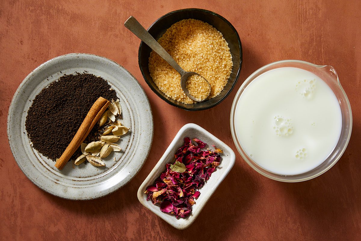 A top view of ingredients in bowls: ground tea leaves, cinnamon sticks, cardamom pods on a plate, brown sugar in a bowl with a spoon, dried rose petals, and a measuring cup of milk, all on a brown surface.