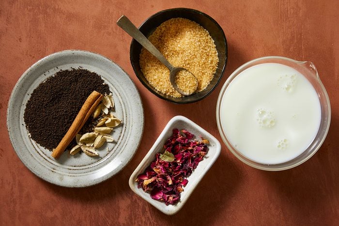 A top view of ingredients in bowls: ground tea leaves, cinnamon sticks, cardamom pods on a plate, brown sugar in a bowl with a spoon, dried rose petals, and a measuring cup of milk, all on a brown surface.