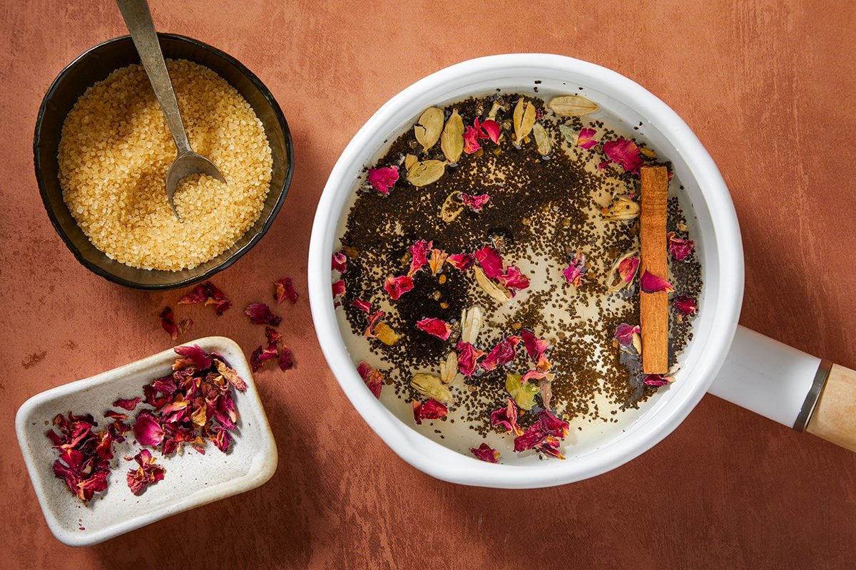 A saucepan filled with milk, loose tea leaves, rose petals, cardamom pods, and a cinnamon stick. Beside it are a bowl of brown sugar with a spoon and a small dish with dried rose petals on a brown surface.