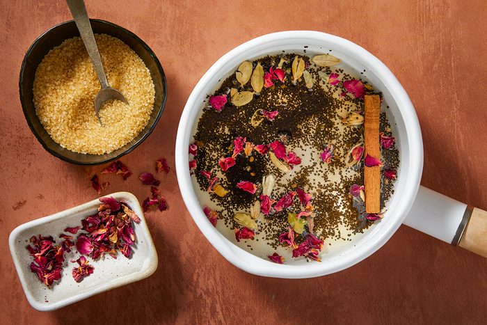 A saucepan filled with milk, loose tea leaves, rose petals, cardamom pods, and a cinnamon stick. Beside it are a bowl of brown sugar with a spoon and a small dish with dried rose petals on a brown surface.