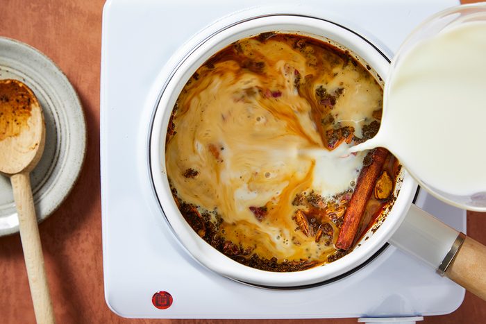 A pot of spiced tea with visible cinnamon sticks simmers on a stovetop as milk is being poured in, creating swirling patterns. A wooden spoon rests on a plate nearby.