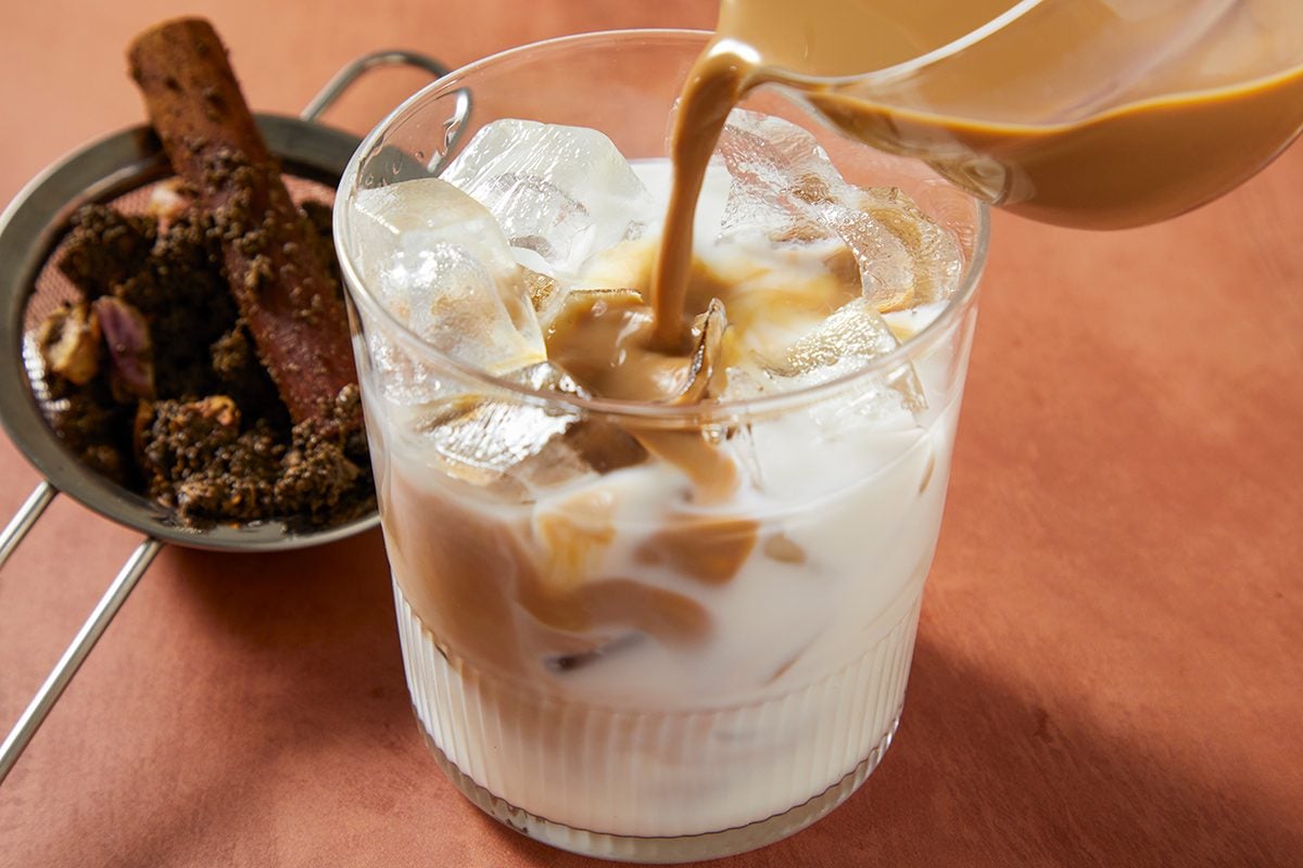 A glass of iced milk is being poured with brown chai tea. Next to the glass is a strainer containing loose chai spices and a cinnamon stick, all placed on a brown surface.