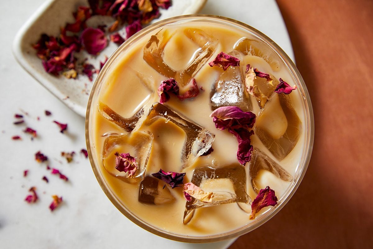A glass of iced milk tea with ice cubes and dried rose petals on top, set on a marble surface with rose petals scattered nearby.