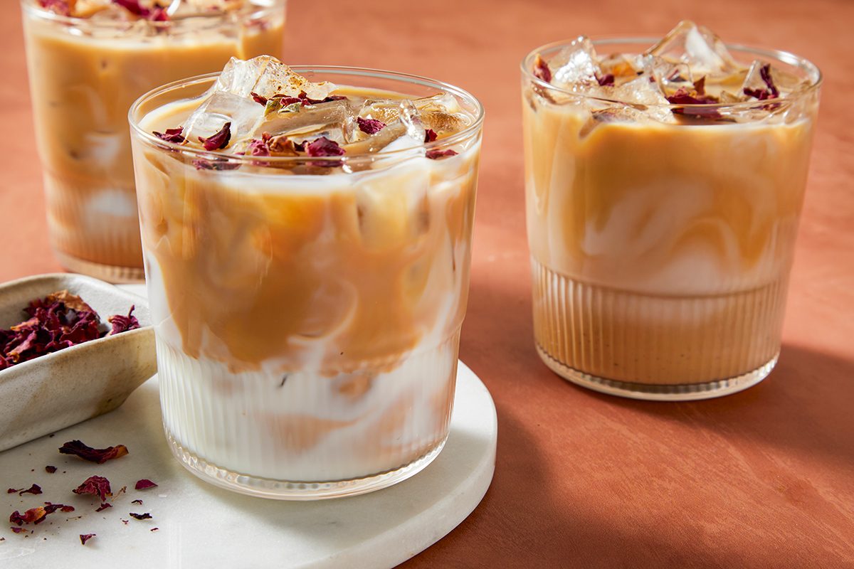 Three glasses of iced coffee with milk sit on a light marble surface. The drinks have a swirled appearance and are topped with ice and dried rose petals, against a warm, brown background.