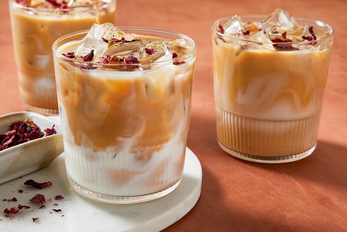 Three glasses of iced coffee with milk sit on a light marble surface. The drinks have a swirled appearance and are topped with ice and dried rose petals, against a warm, brown background.