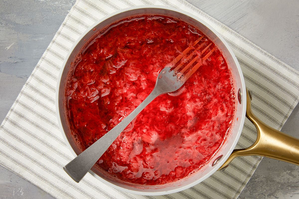 Mashing softened strawberries in the saucepan to break down the fruit into a chunky compote.