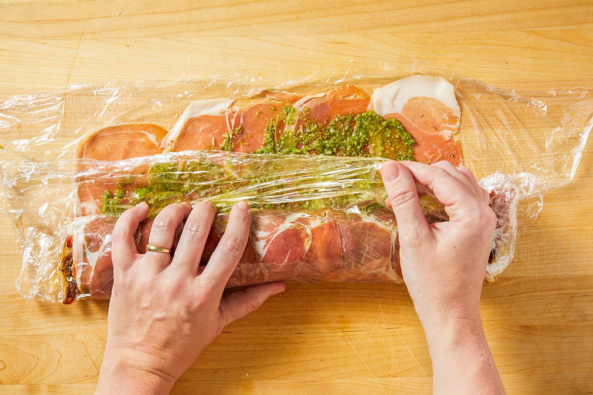 The layered ingredients are tightly rolled into a log using plastic wrap to shape the Italian sushi.