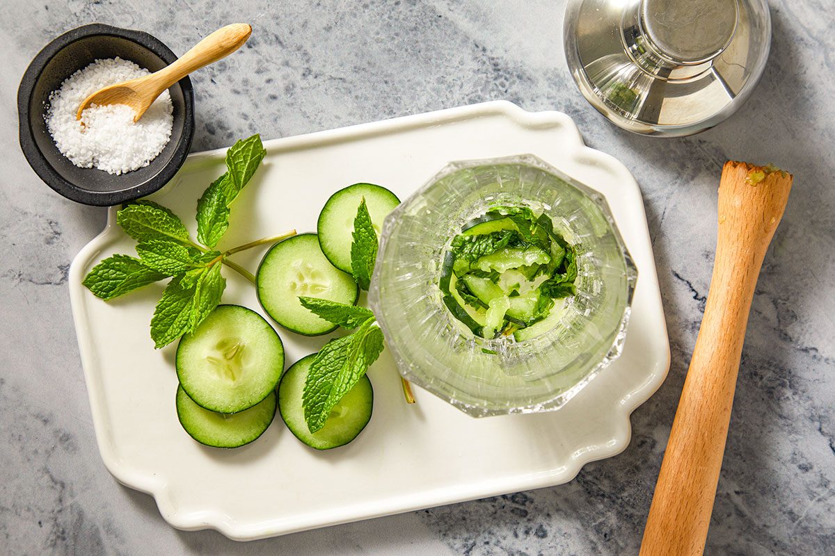 A horizontal overhead image shows cucumber slices and fresh mint being muddled with salt in a mixing vessel alongside a citrus juicer and bar tools.