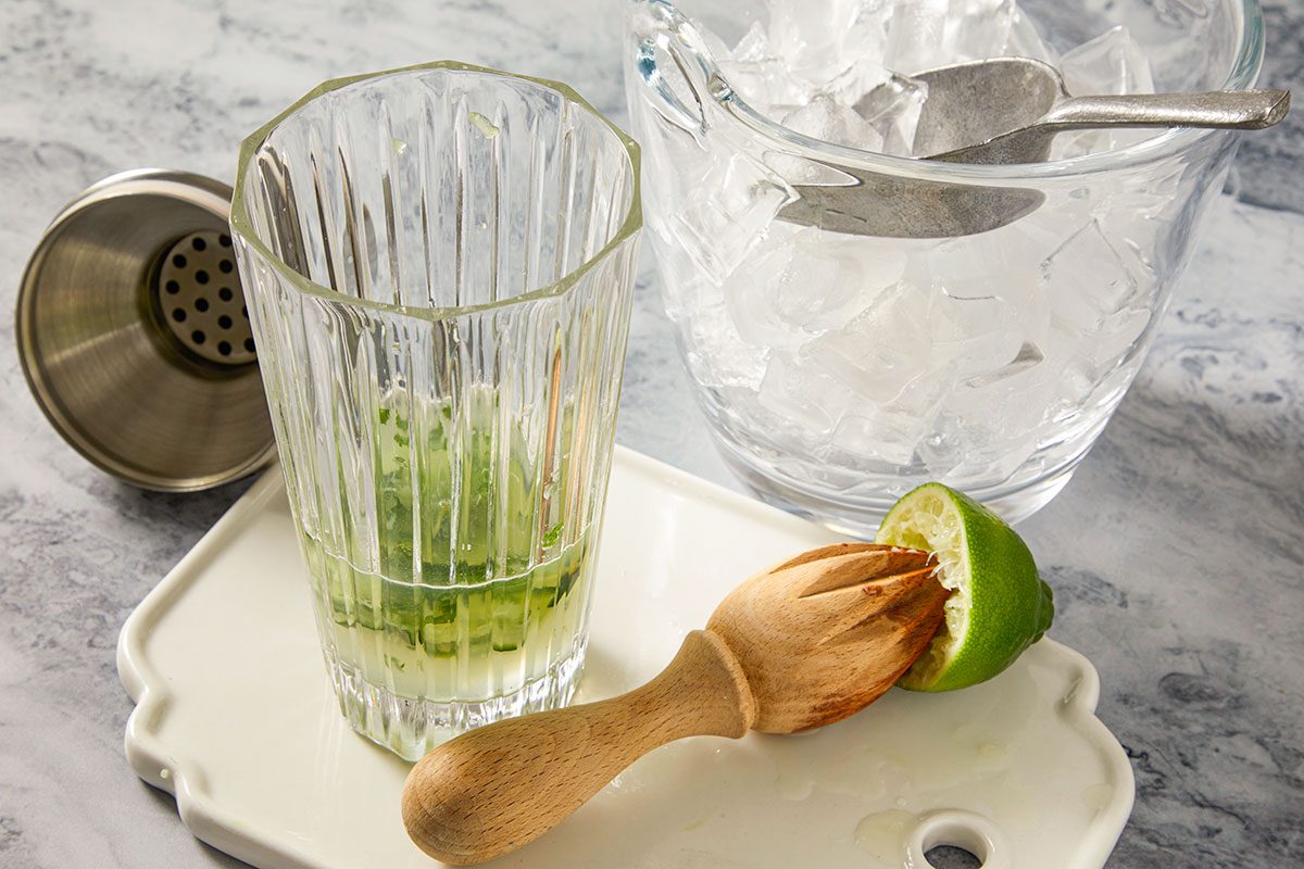 A horizontal overhead image shows muddled cucumber and mint in a glass with lime juice being added, alongside ice and cocktail tools.