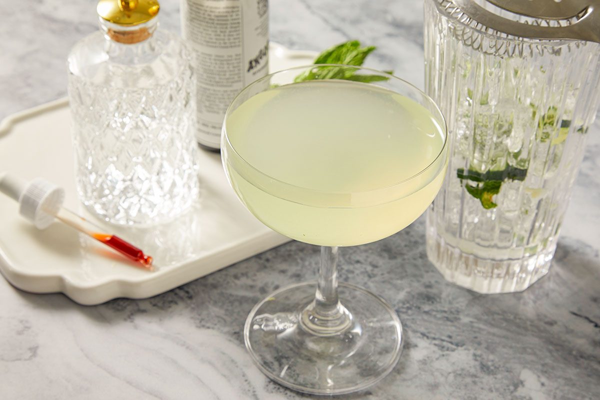 A horizontal image shows a pale green cocktail strained into a coupe glass with bar tools and ingredients in the background.