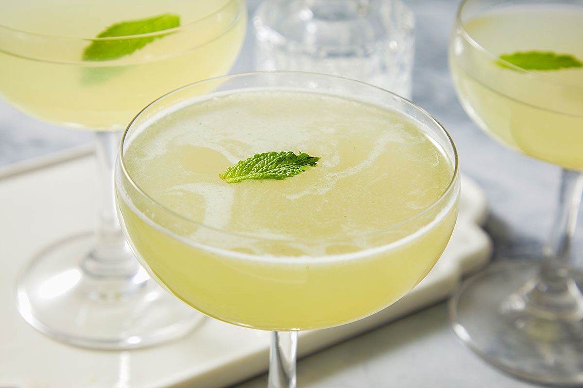 A horizontal image shows several coupe glasses filled with the pale green cocktail, each garnished with mint leaves.