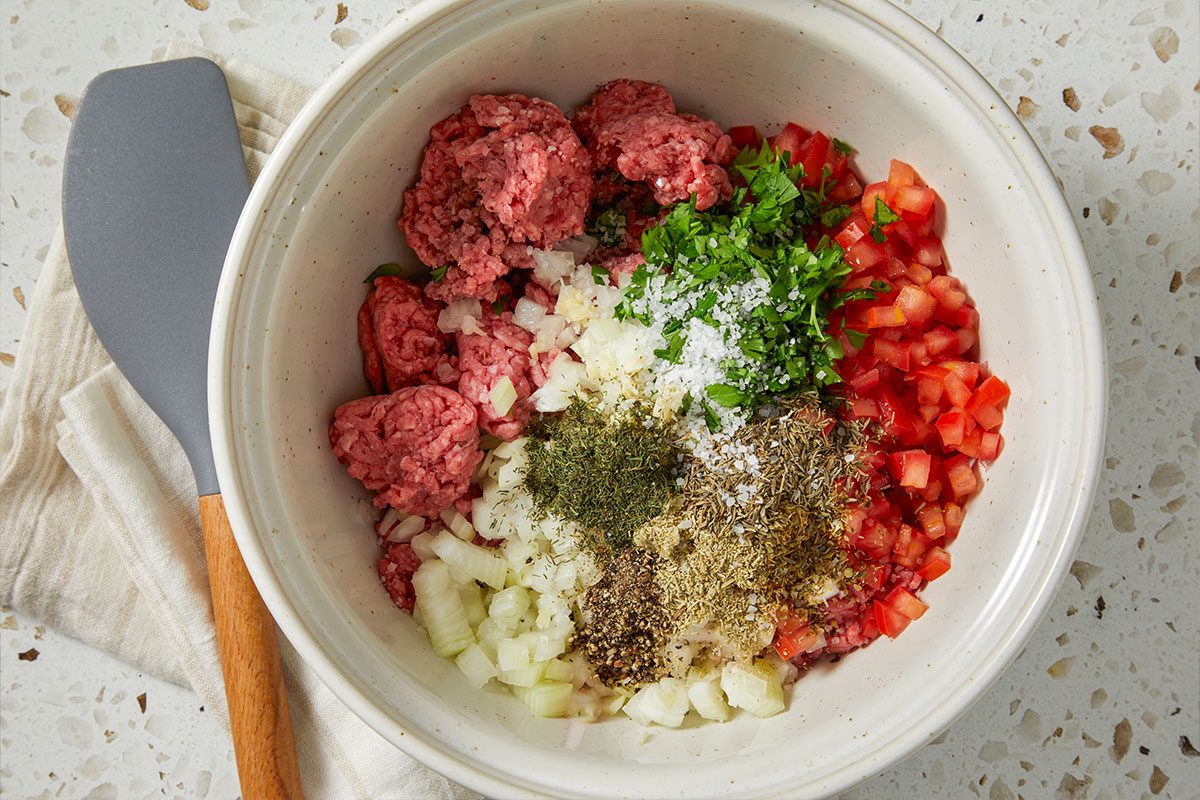 Overhead horizontal image of a mixing bowl filled with the recipe's ingredients. The visible ingredients include ground meat, diced tomatoes and onions, various spices and herbs, and salt. A gray spatula with a wooden handle is on the left side of the bowl.