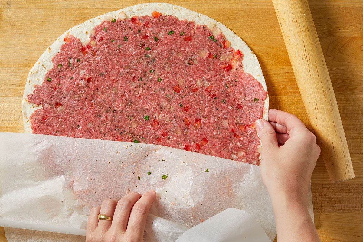 Overhead horizontal image of a tortilla being prepared. A ground meat mixture has been added to half of the tortilla, and a hand is seen layering a piece of parchment paper over the top. A rolling pin rests on the side of the wooden cutting board.