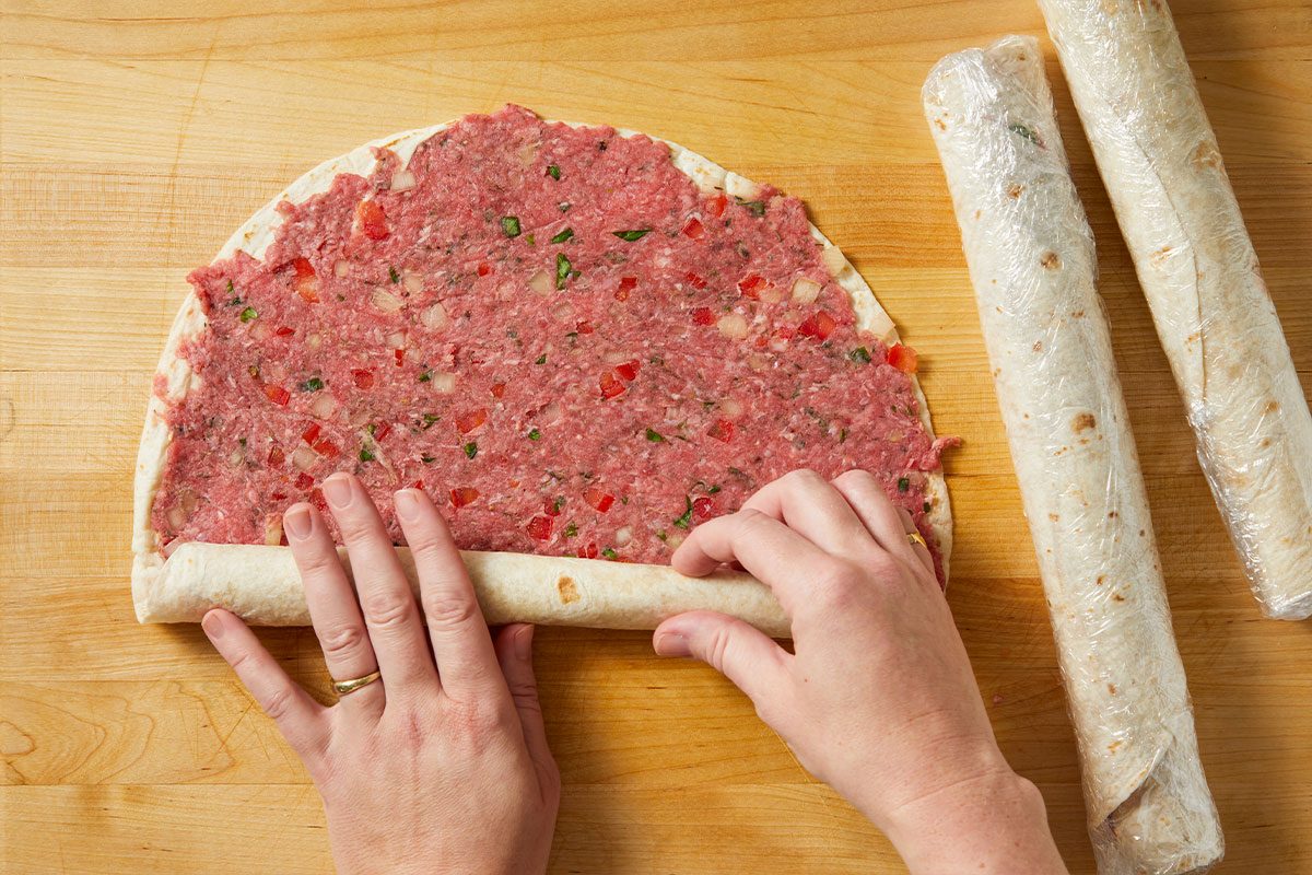 Overhead horizontal image of the step-by-step process of rolling up a tortilla filled with ground meat, diced tomatoes, and onions. Two rolled tortillas are visible, wrapped in plastic, to the side.