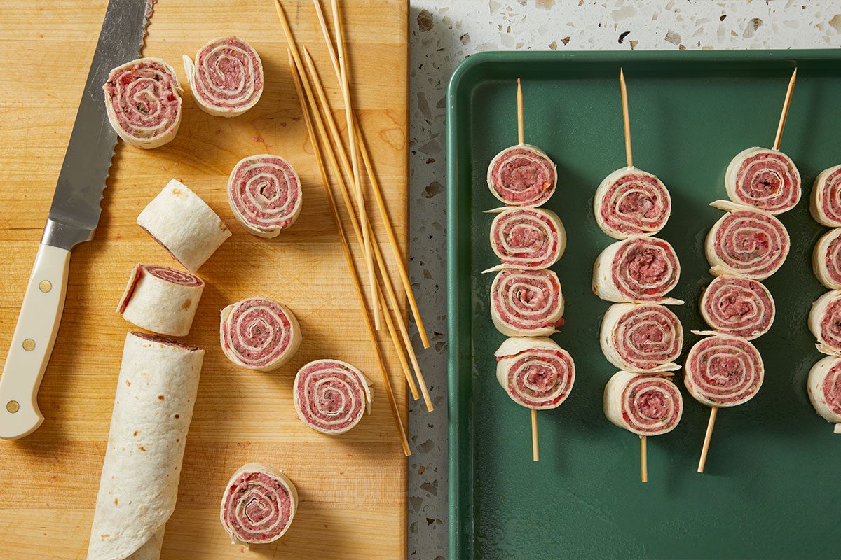 Overhead horizontal image of pinwheel appetizers. Some of the sliced pinwheels are on a wooden cutting board, next to a long knife and wooden skewers, and others are arranged on a green baking sheet, skewered three at a time. The filling appears to include meat and possibly herbs, all wrapped in a white tortilla.