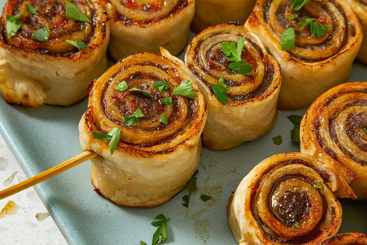 Eye Level horizontal image of savory roll ups, with a wooden skewer through one. The filling is visible through the spirals of the dough, and parsley sprigs are scattered on top.