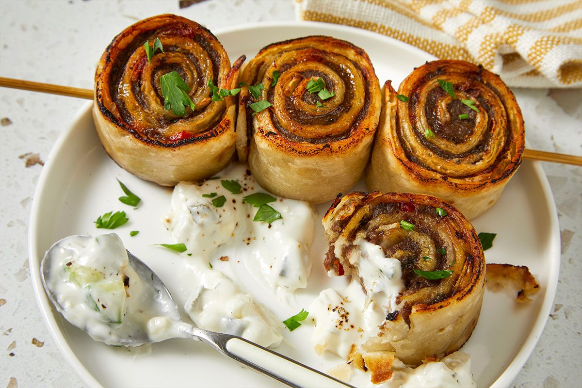 Eye Level horizontal image of four rolls on a white plate, three on a wooden skewer. The rolls are garnished with parsley and accompanied by a creamy sauce; a spoon holds a portion of the sauce with visible cucumber chunks.