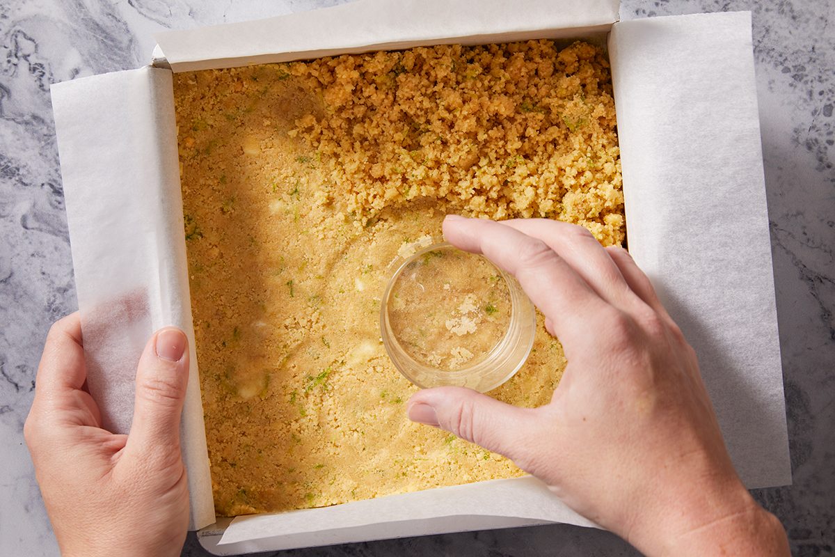 A person presses a crumbly crust mixture into a square baking pan lined with parchment paper, using the bottom of a glass to flatten the crust evenly.