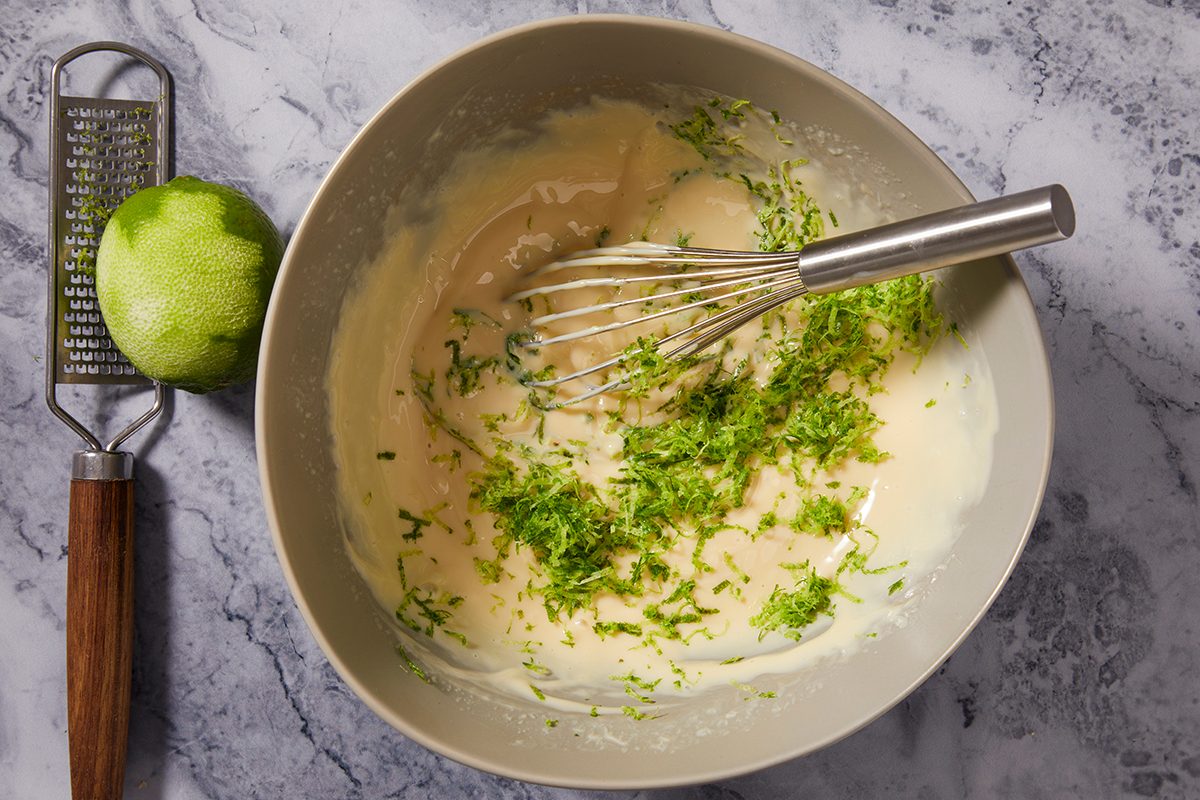 A mixing bowl with batter, a metal whisk, and freshly grated lime zest on top. Beside the bowl are a whole lime and a grater on a gray marble surface.