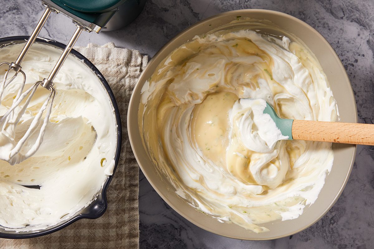 A mixing bowl filled with creamy batter being stirred with a rubber spatula next to a hand mixer with beaters covered in batter, sitting on a checkered cloth on a marble countertop.