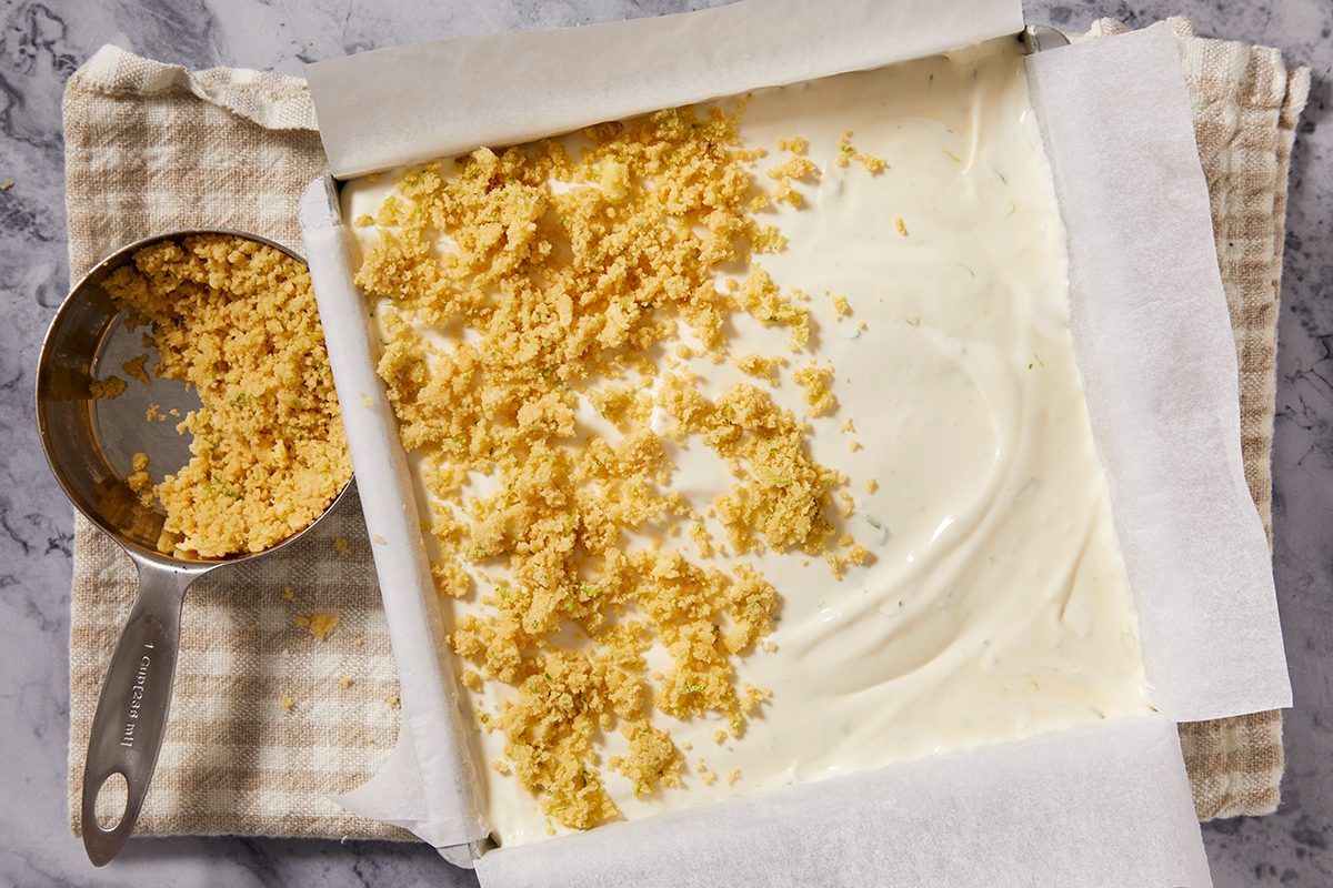 A square baking pan lined with parchment paper is filled with a creamy white mixture, partially topped with crumbled yellow dough. A metal measuring cup with more crumbs rests beside the pan on a cloth.