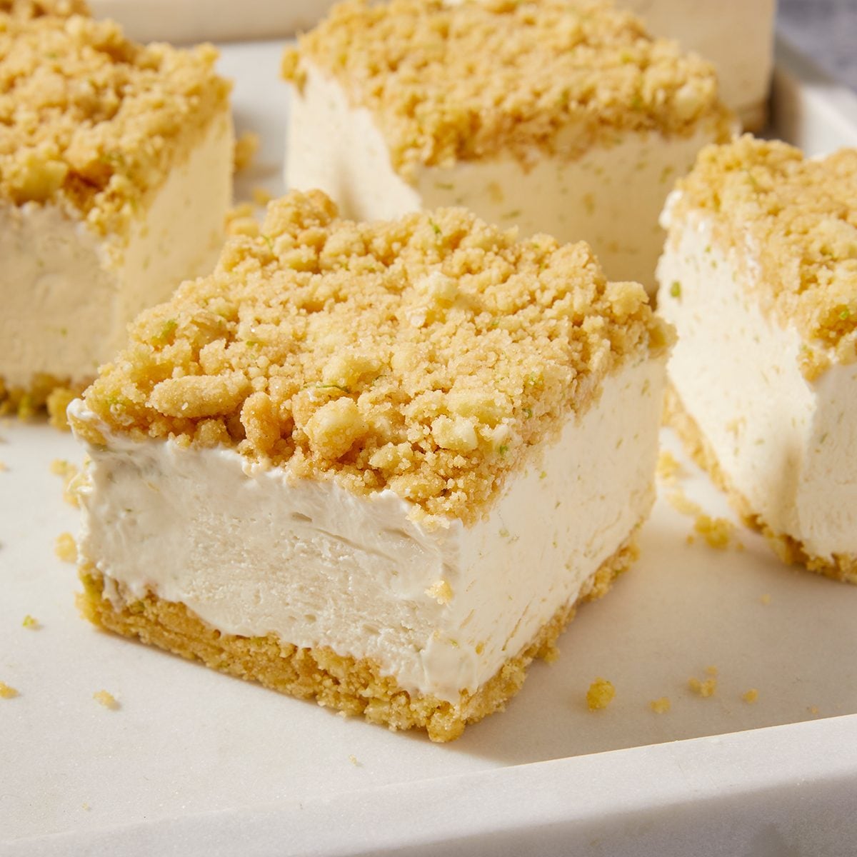Three thick, square ice cream sandwiches are shown, made with a creamy vanilla ice cream filling between two layers of golden, crumbly cookie crust. The sandwiches are displayed on a white surface.