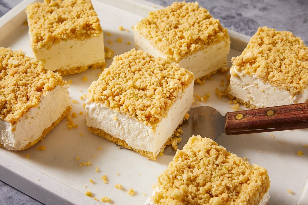 Six square pieces of crumbly cheesecake bars with a golden streusel topping and crust are arranged on a white tray; one piece is being lifted with a metal spatula with a wooden handle.
