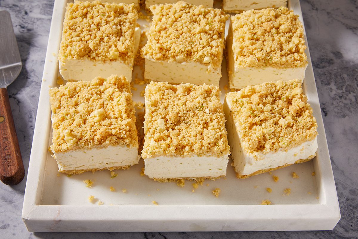 Nine square pieces of crumb-topped cheesecake bars arranged neatly on a white rectangular tray, with a knife visible to the side on a marble surface.