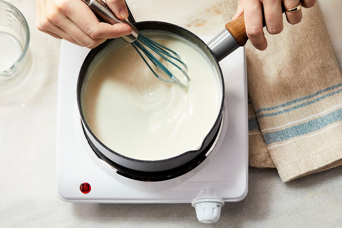 A person is whisking a creamy white liquid in a small saucepan on an electric stovetop, with a tan towel and glass nearby.
