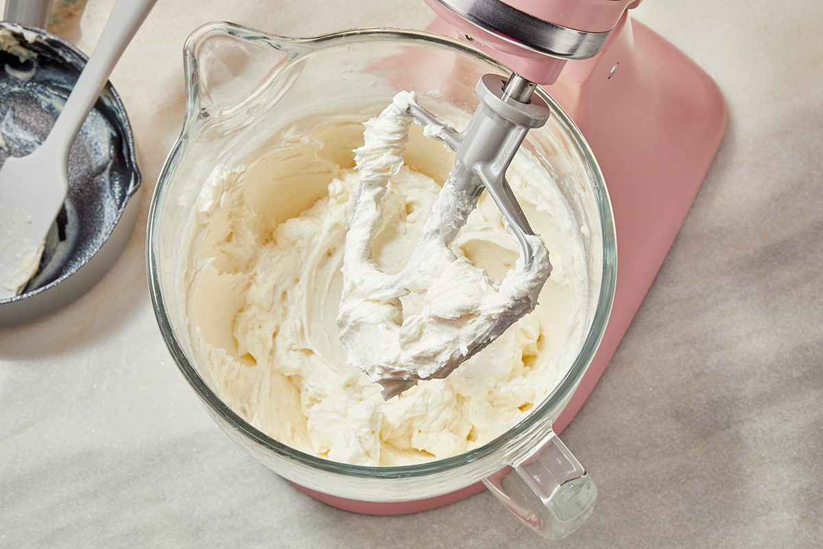 A glass mixing bowl attached to a pink stand mixer contains whipped cream or frosting, with a paddle attachment coated in the creamy mixture. A spatula rests in a bowl nearby on a marble countertop.