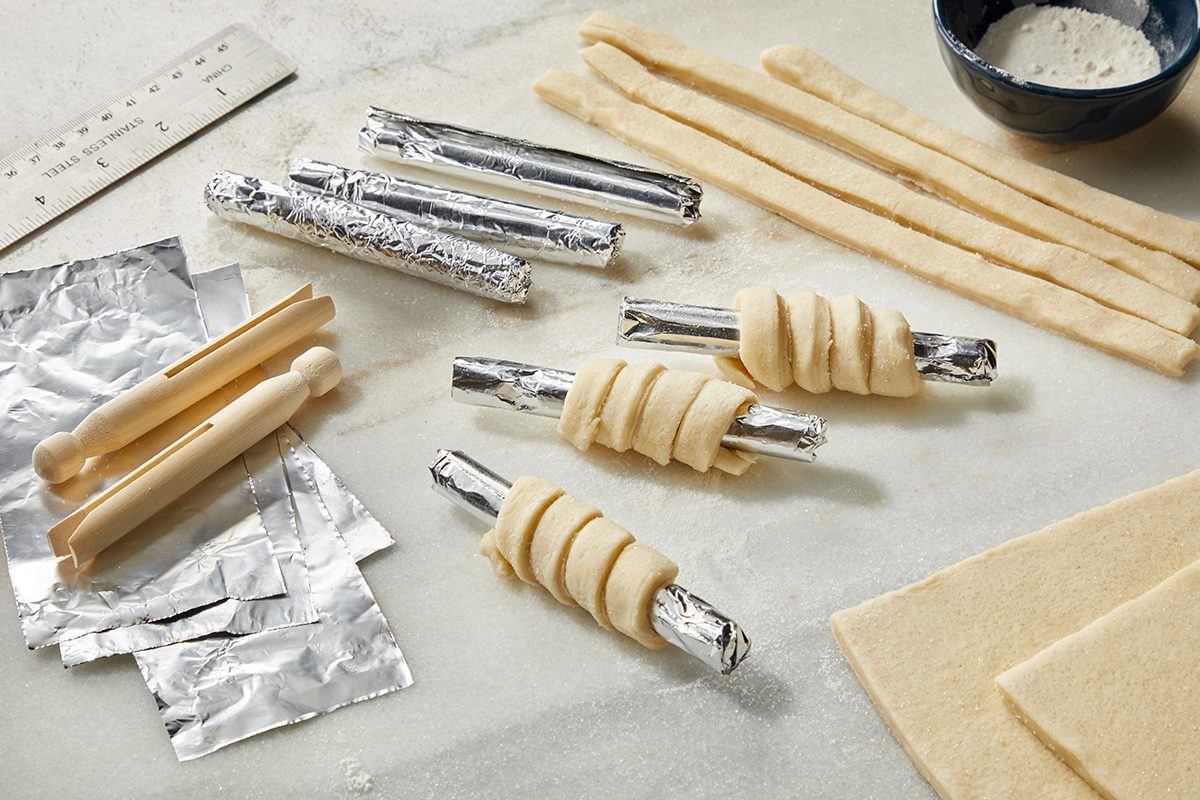 Puff pastry strips are wrapped around foil-covered tubes on a countertop, with extra dough strips, a ruler, a bowl with white contents, and pieces of foil visible nearby.