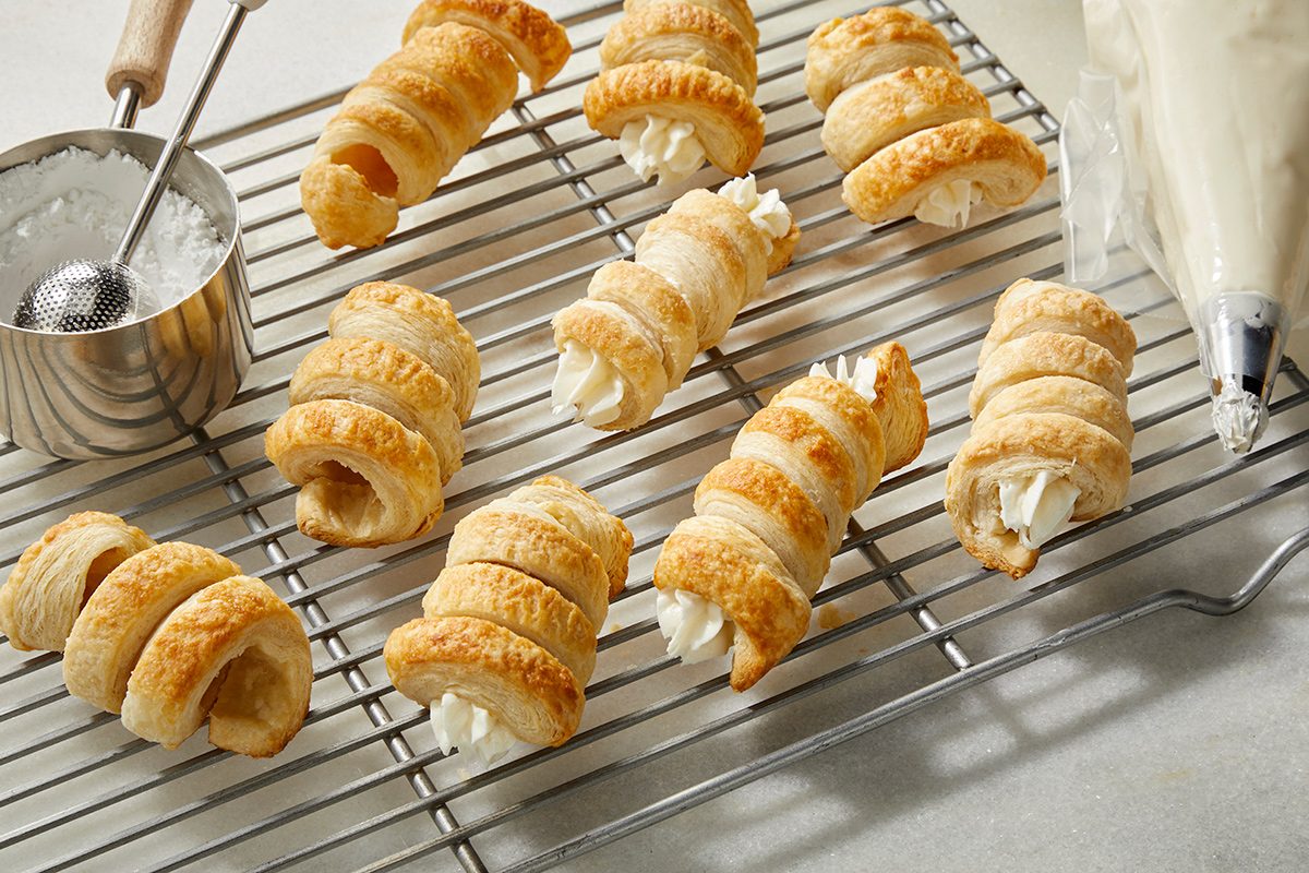 Golden puff pastry horns filled with a creamy white filling are cooling on a wire rack. A metal cup, a spoon, and a piping bag filled with cream are nearby on a light-colored surface.