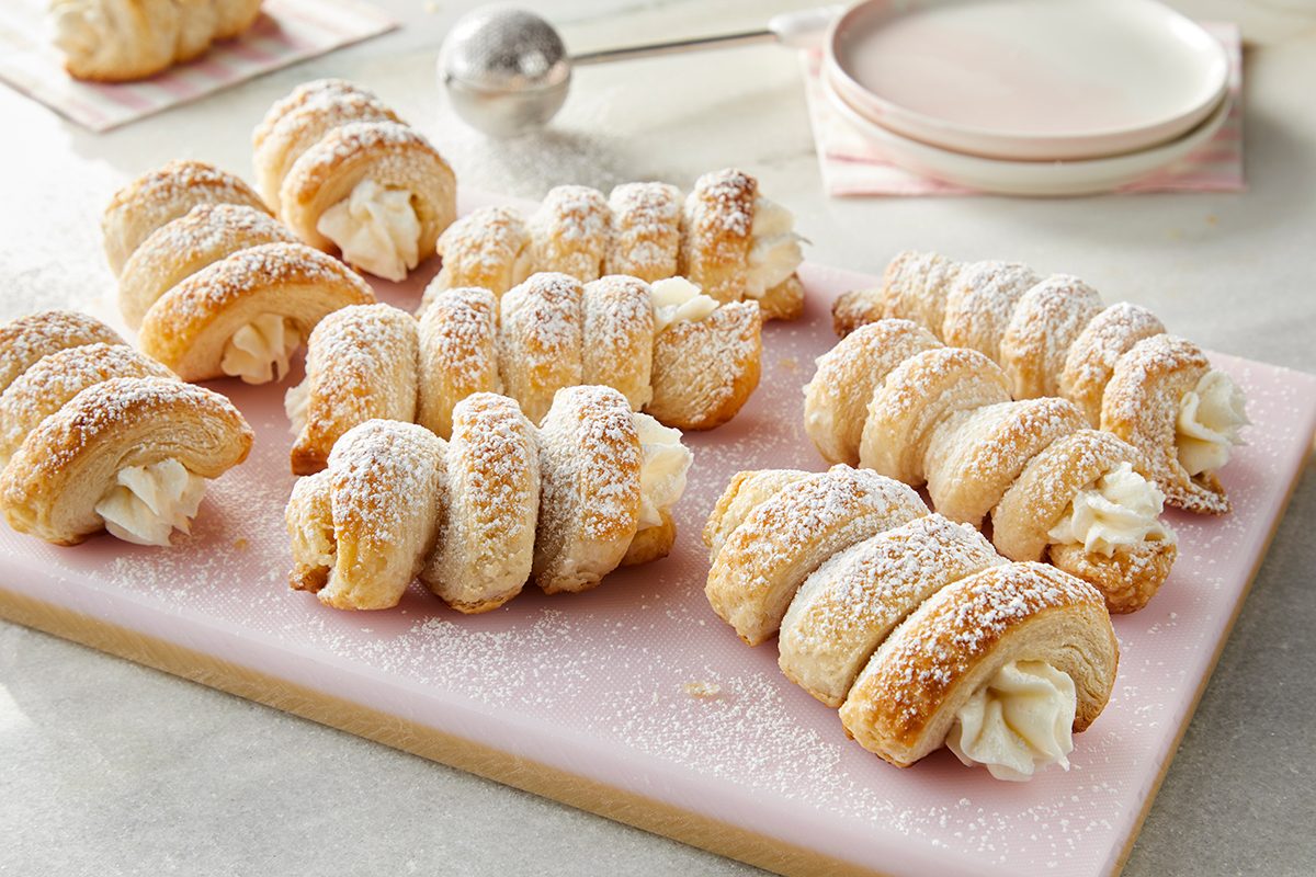 A group of cream-filled pastry horns dusted with powdered sugar are arranged on a light pink cutting board, with plates and a sugar sifter in the background.