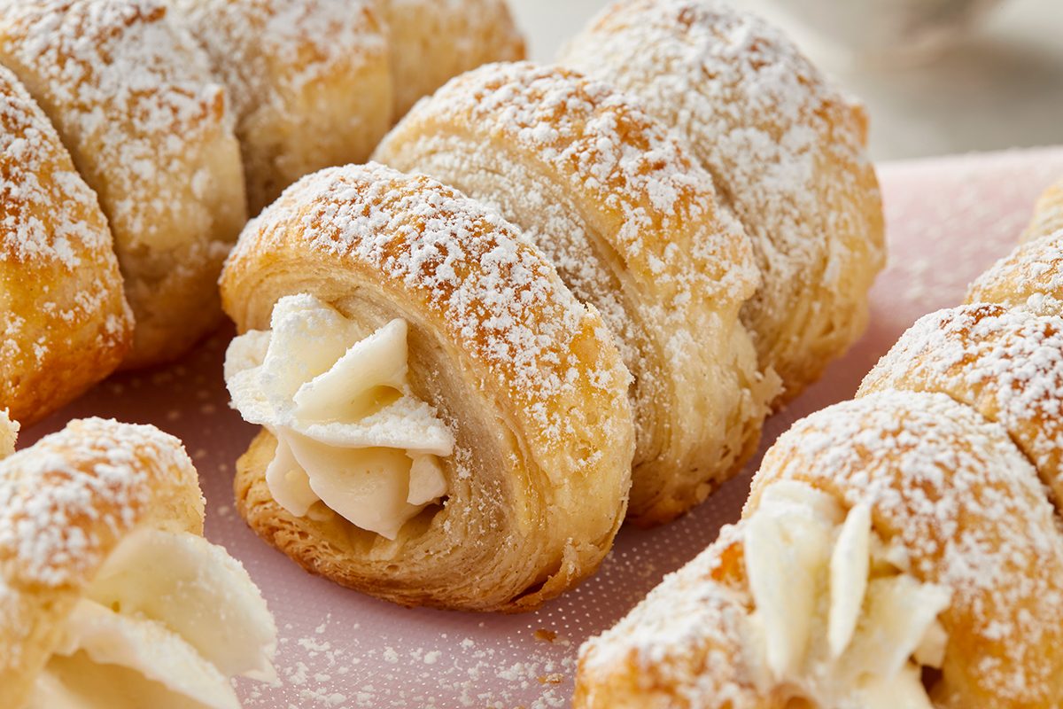Close-up of golden, flaky pastry horns filled with cream and dusted with powdered sugar, arranged on a light pink surface.