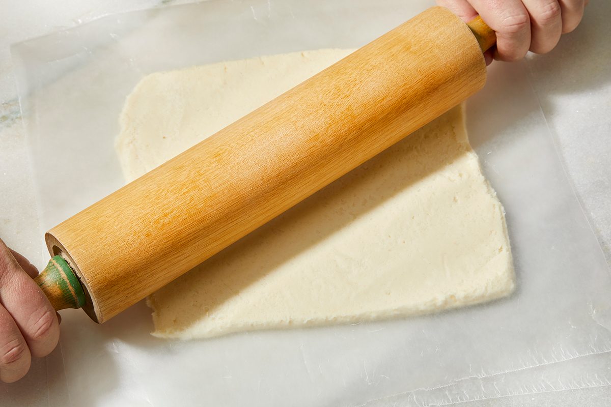 A person uses a wooden rolling pin to flatten dough between two sheets of parchment paper on a white surface.