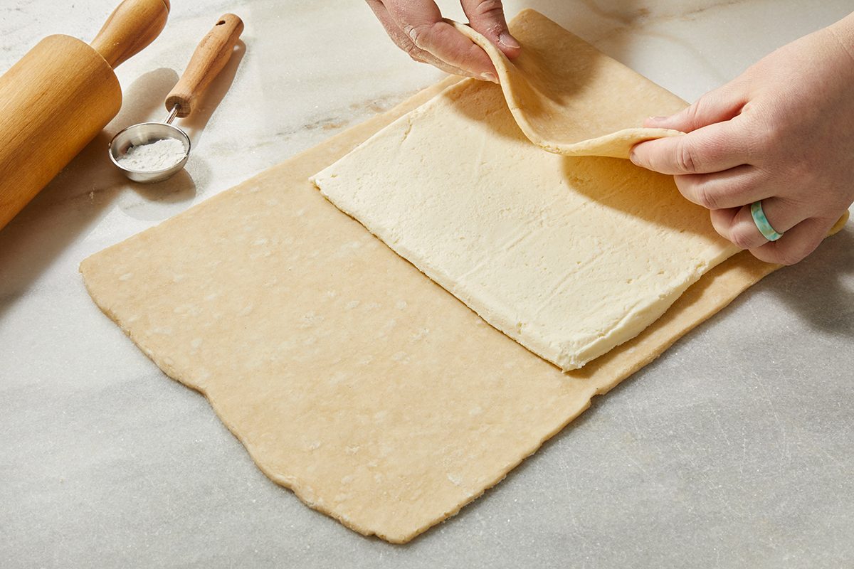 Hands folding a sheet of dough over a square of butter on a marble surface, with a rolling pin and small dish nearby, preparing laminated dough for baking.