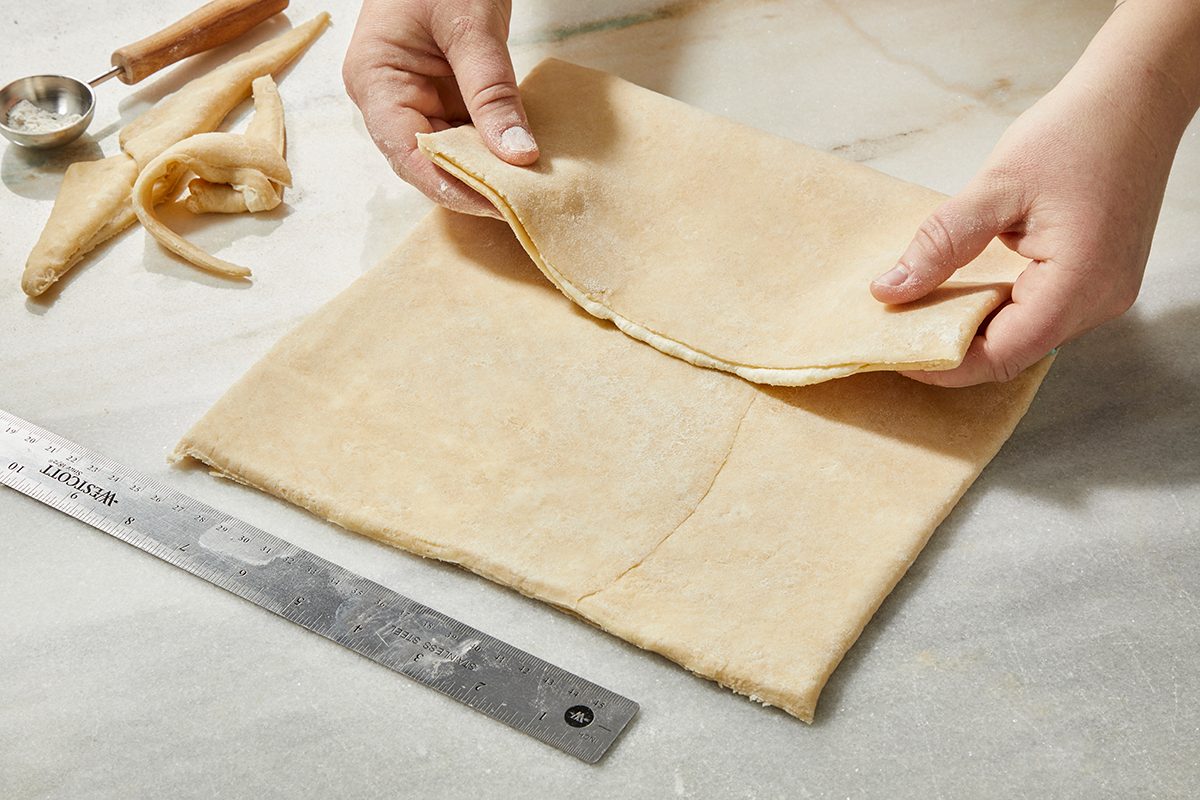 Hands folding a square sheet of dough on a marble surface, with a ruler, pastry cutter, and some dough scraps nearby.