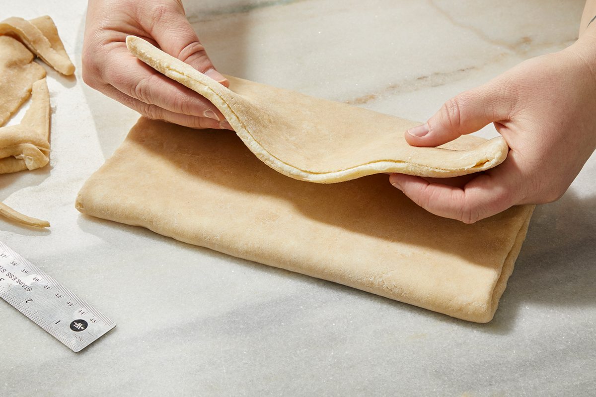 Two hands fold a sheet of dough on a marble surface, with a metal ruler nearby.