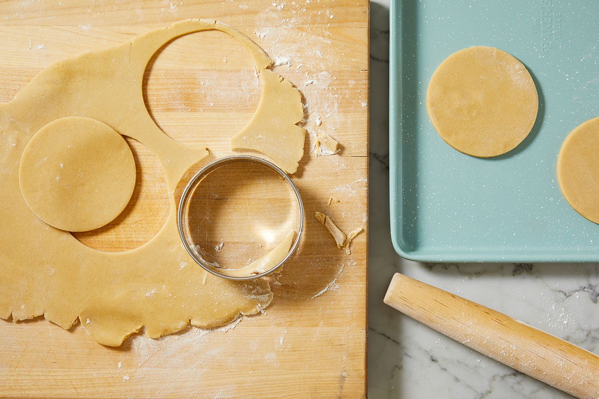 A horizontal overhead image shows rolled cookie dough being cut into rounds on a floured surface with a biscuit cutter and rolling pin.