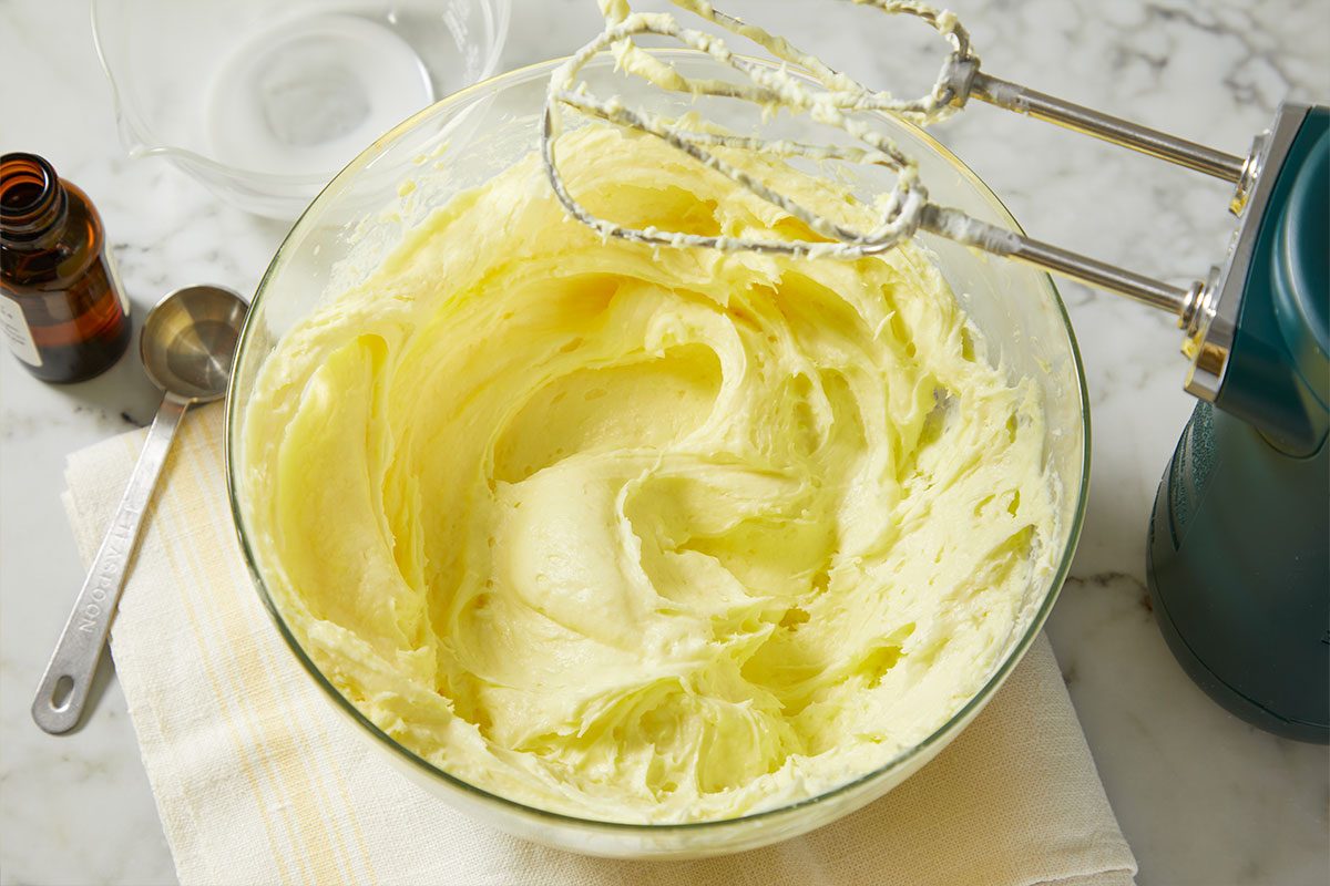 A horizontal overhead image shows a creamy lemon mascarpone frosting being mixed in a bowl with a hand mixer.