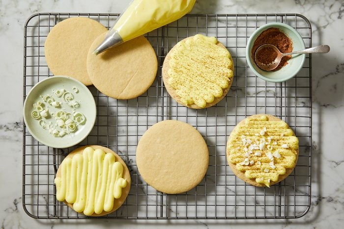 A horizontal overhead image shows baked cookies on a cooling rack as frosting is piped onto the tops with a piping bag, with cocoa nearby.