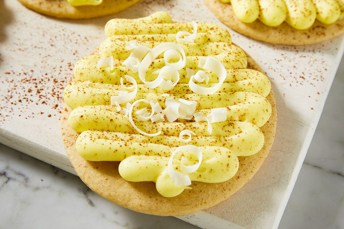 A horizontal close-up image shows a single frosted lemon cookie topped with piped frosting and white chocolate curls.