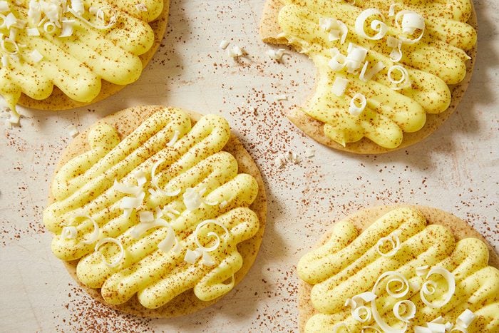 A horizontal image shows several frosted lemon tiramisu cookies arranged together with visible frosting ridges and chocolate garnish.