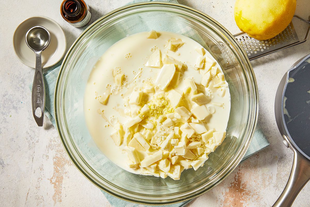 Overhead horizontal image of heavy cream and chopped white chocolate in a bowl with lemon zest and extract nearby for truffle base.