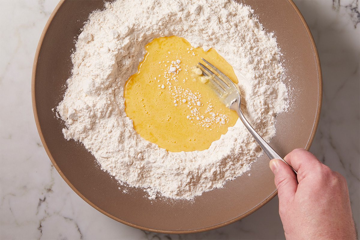 Overhead step-by-step preparation image showing eggs being whisked into a mound of all-purpose flour to create fresh pasta dough. The flour well technique is used on a plate over a marble surface, illustrating the first stage of homemade lobster ravioli pasta dough preparation.