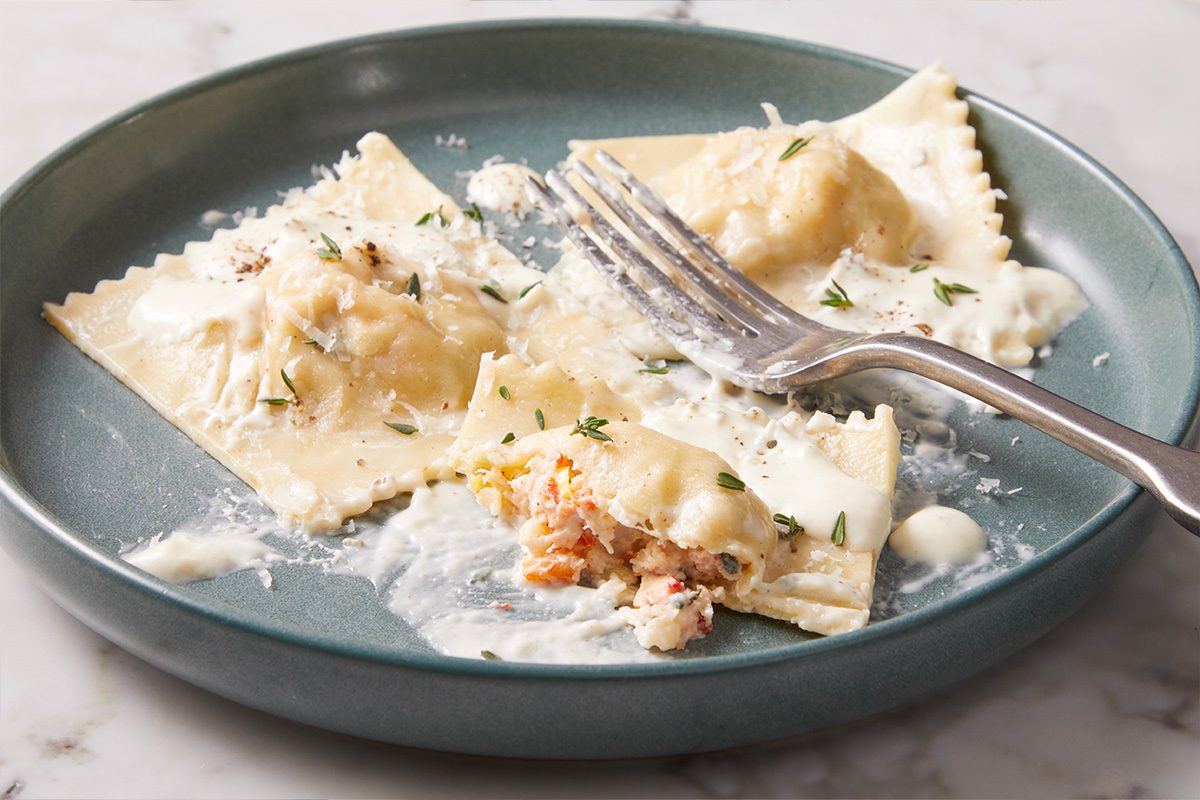 Close-up of lobster ravioli topped with grated Parmesan and creamy sauce on a teal plate, with a fork lifting a ravioli piece to reveal the lobster filling.