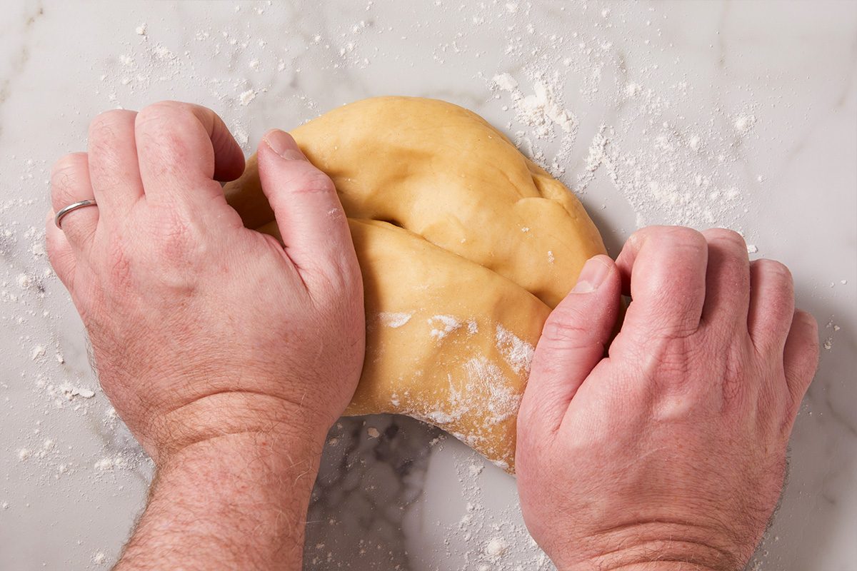 Hands kneading fresh pasta dough on a lightly floured marble surface during homemade ravioli preparation. The smooth dough is being folded and pressed to develop gluten structure for tender ravioli pasta.