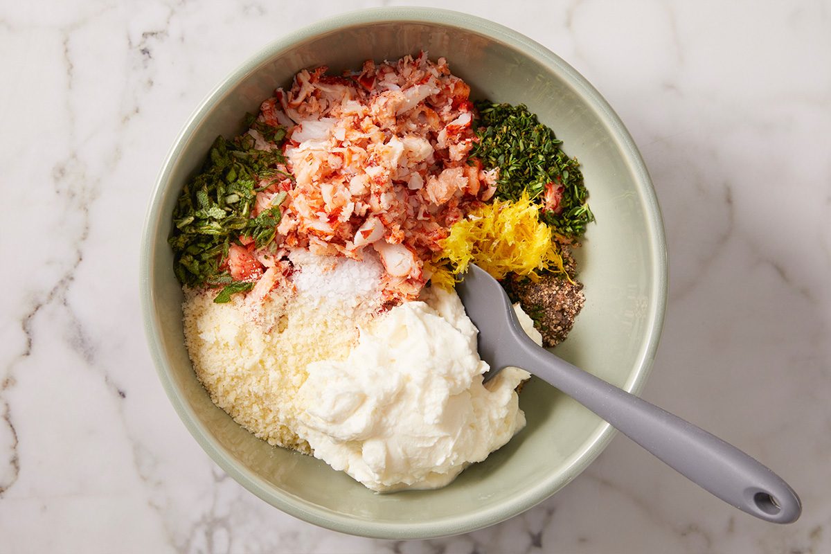 Overhead view of lobster ravioli filling ingredients in a bowl including chopped cooked lobster meat, ricotta cheese, grated Parmesan, lemon zest and fresh herbs being mixed together to create a creamy seafood filling.